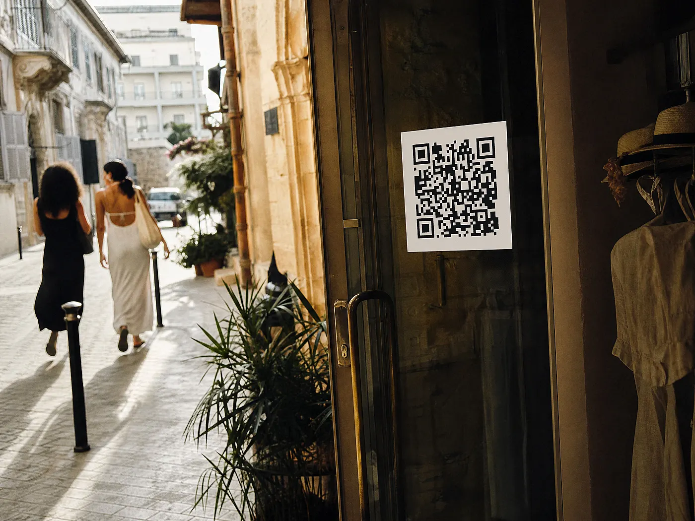 Bir butiğin cam kapısında QR etiketi. Yanındaki telefon, aynı QR'ın açtığı sayfanın Ekim'de Yaz indirimi, Kasım'da Yeni sezon olduğunu gösteriyor.